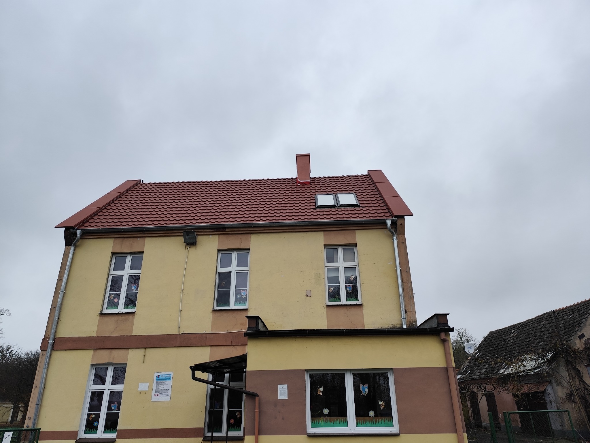 Dwupiętrowy budynek z czerwoną dachówką i oknami ozdobionymi naklejkami, widziany z dołu na tle pochmurnego nieba. Widoczny komin i fragment sąsiedniego budynku.