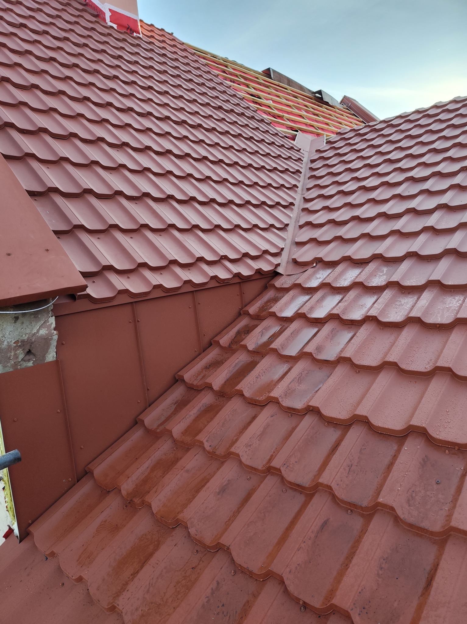 Czerwona blachodachówka na skośnym dachu, widoczne krople wody i fragment więźby dachowej. Ujęcie z dołu, podkreślające strukturę pokrycia.