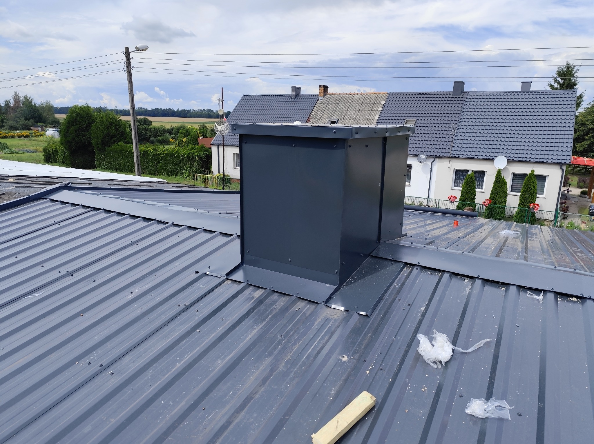 Szary dach z blachy trapezowej z kominem wentylacyjnym na tle zabudowań i błękitnego nieba. Widoczne ślady montażu i akcesoria dekarskie.