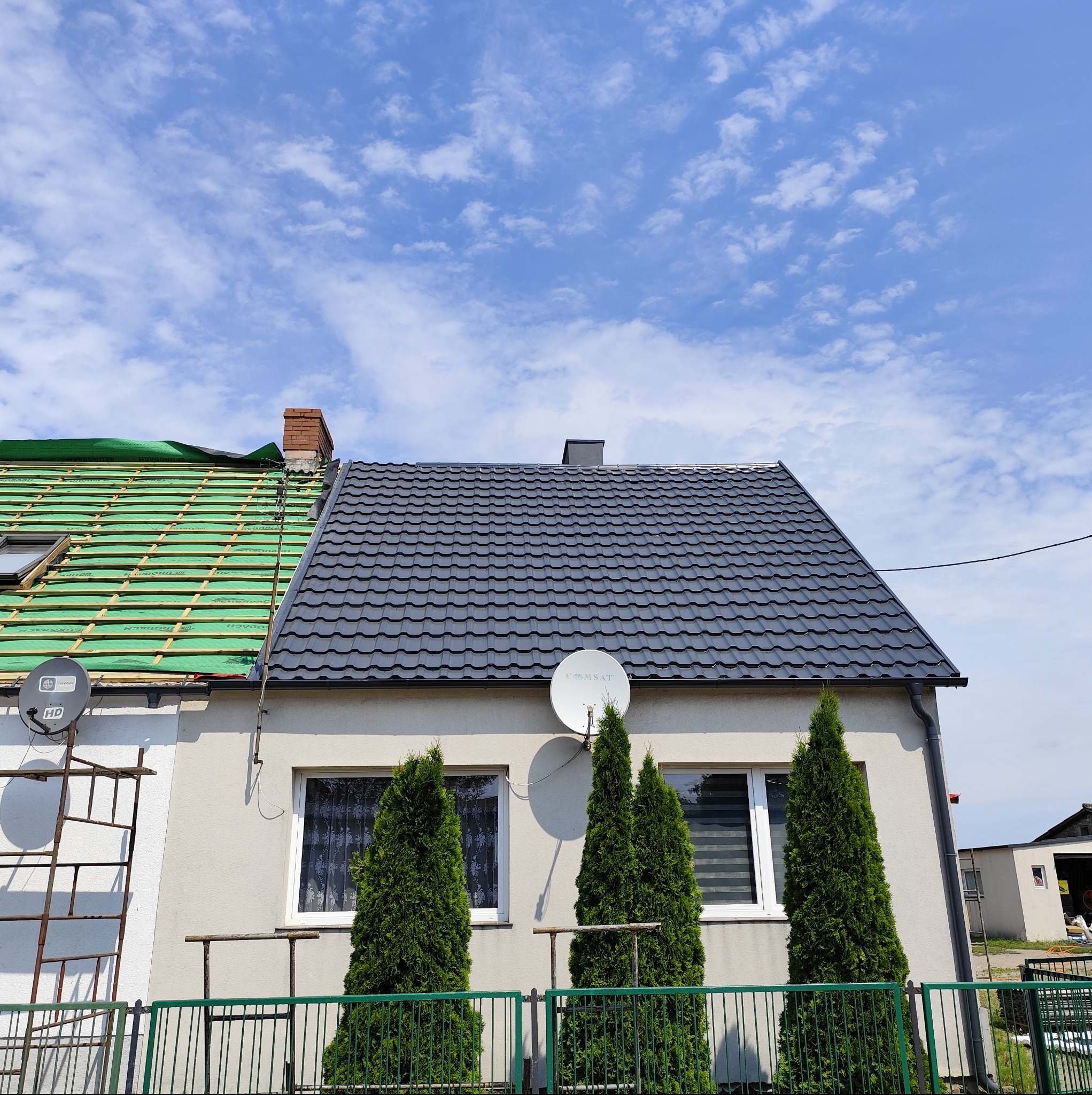 Dom z częściowo wymienionym dachem: połowa pokryta szarą blachodachówką, druga w trakcie prac z zieloną folią. Widoczne rusztowania i trzy tuje przed budynkiem.