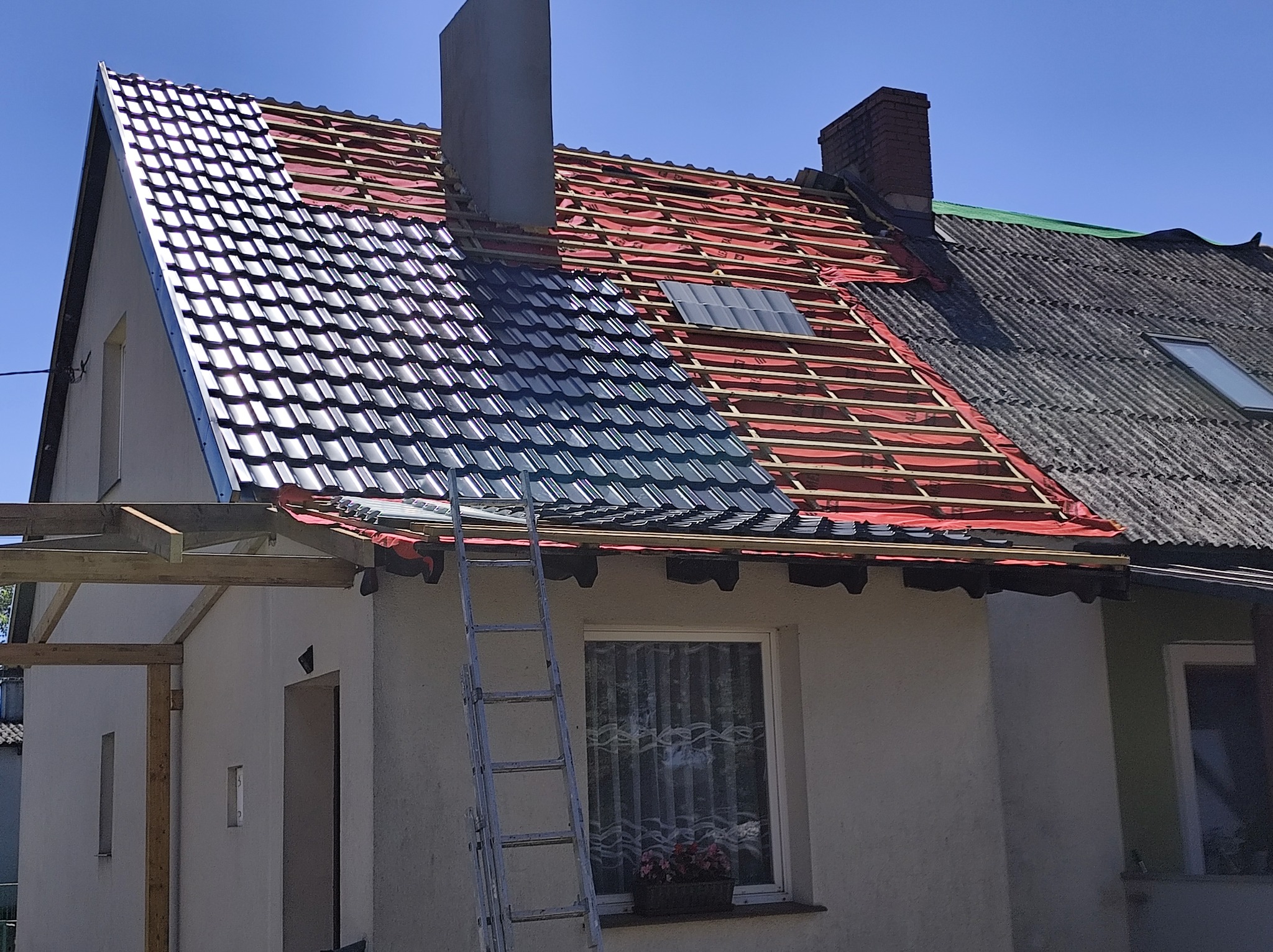 Częściowo wymieniony dach z czarną dachówką, widoczna więźba i komin. Stara część dachu z eternitu. Drabina oparta o ścianę budynku. Słoneczny dzień.