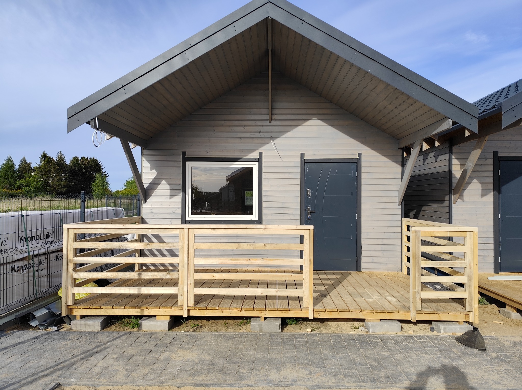 Gotowy, szary dom szkieletowy z drewnianym tarasem w Jagniewicach. Widoczne materiały budowlane w tle. Dach dwuspadowy, okno z białą ramą, szare drzwi.