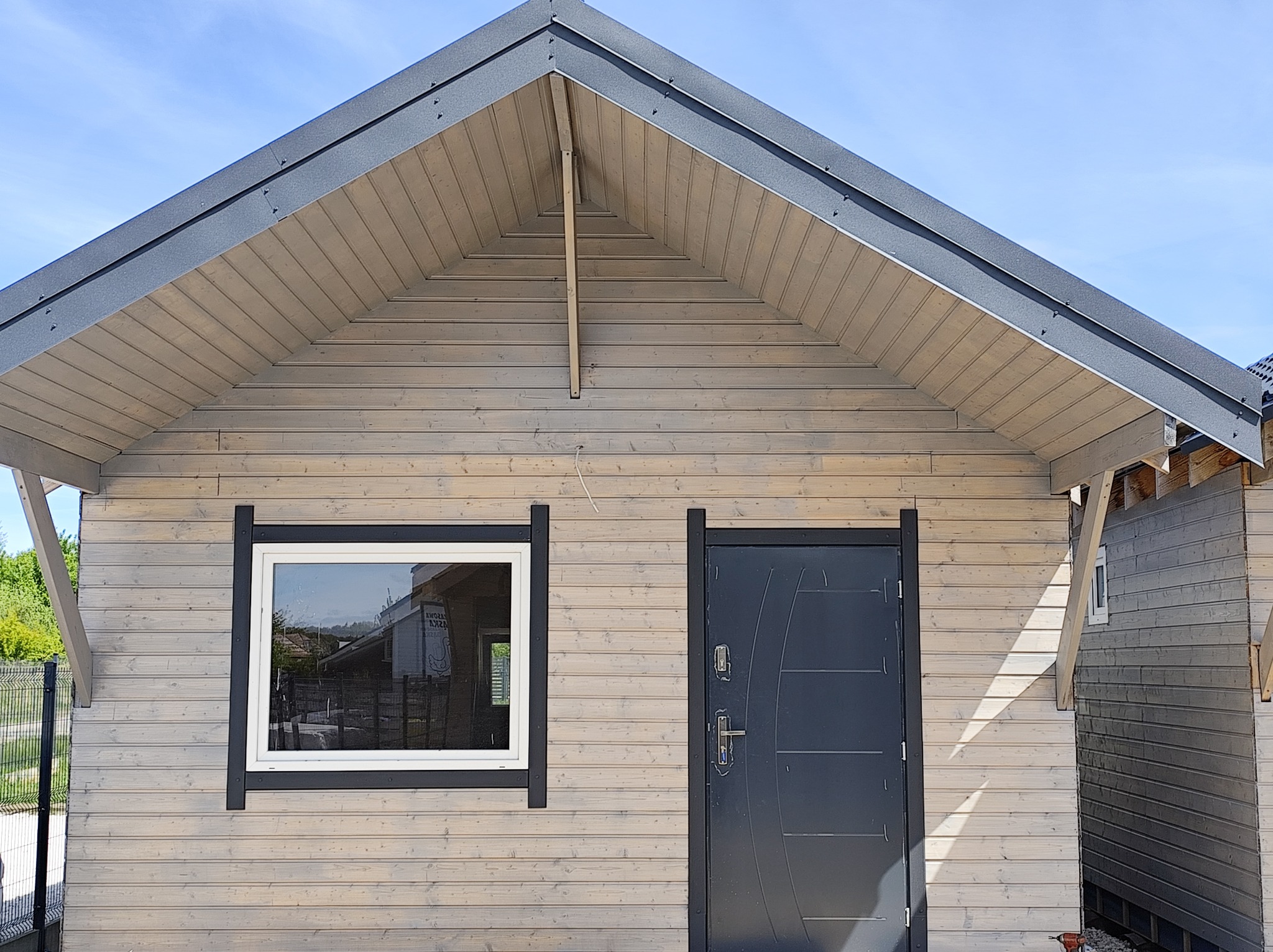 Elewacja domu szkieletowego z jasnego drewna z ciemnymi obramowaniami okna i drzwi. Widoczny fragment dachu i boczna ściana sąsiedniego budynku.