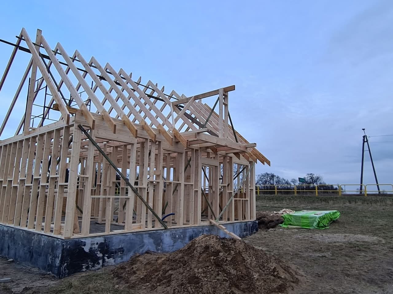 Surowy szkielet domu w trakcie budowy, widoczna drewniana konstrukcja ścian i dachu na fundamencie, z ziemią i zielonym materiałem budowlanym w tle.