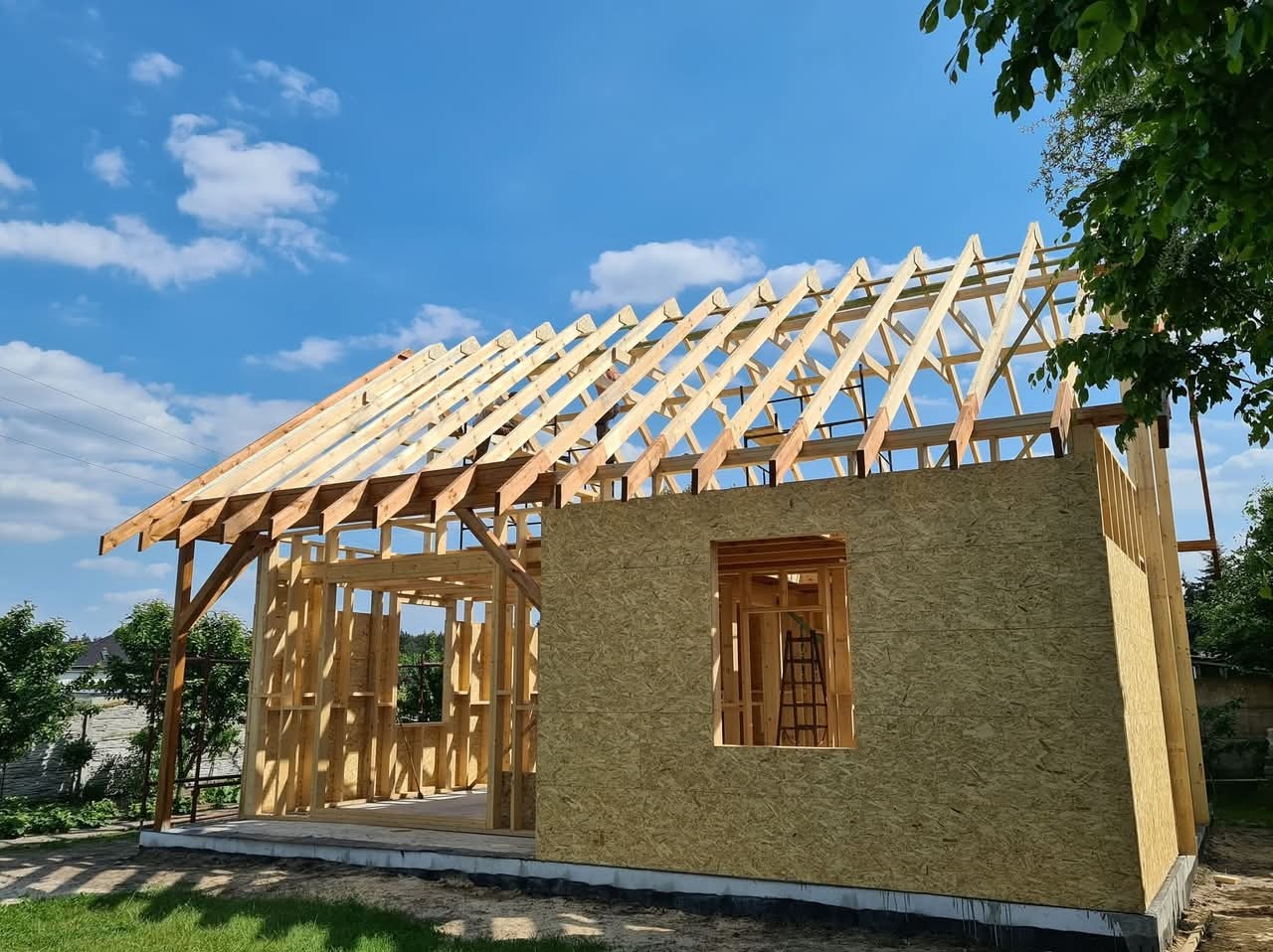 Konstrukcja domu szkieletowego w trakcie budowy, widoczna więźba dachowa i ściany z płyt OSB, błękitne niebo w tle. Ujęcie z poziomu gruntu.