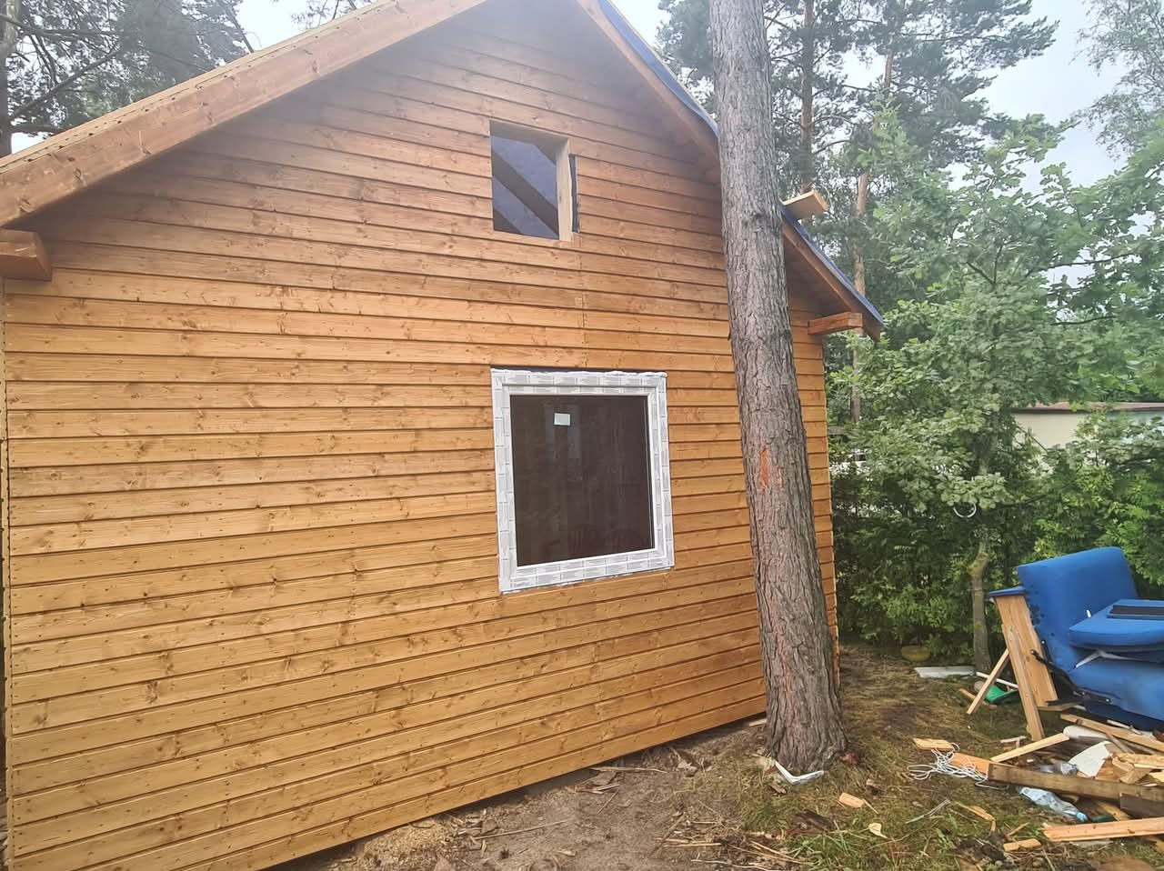 Drewniany dom szkieletowy w trakcie budowy, z widocznymi oknami i deskami elewacyjnymi. Obok domu rośnie drzewo, a w tle zieleń.