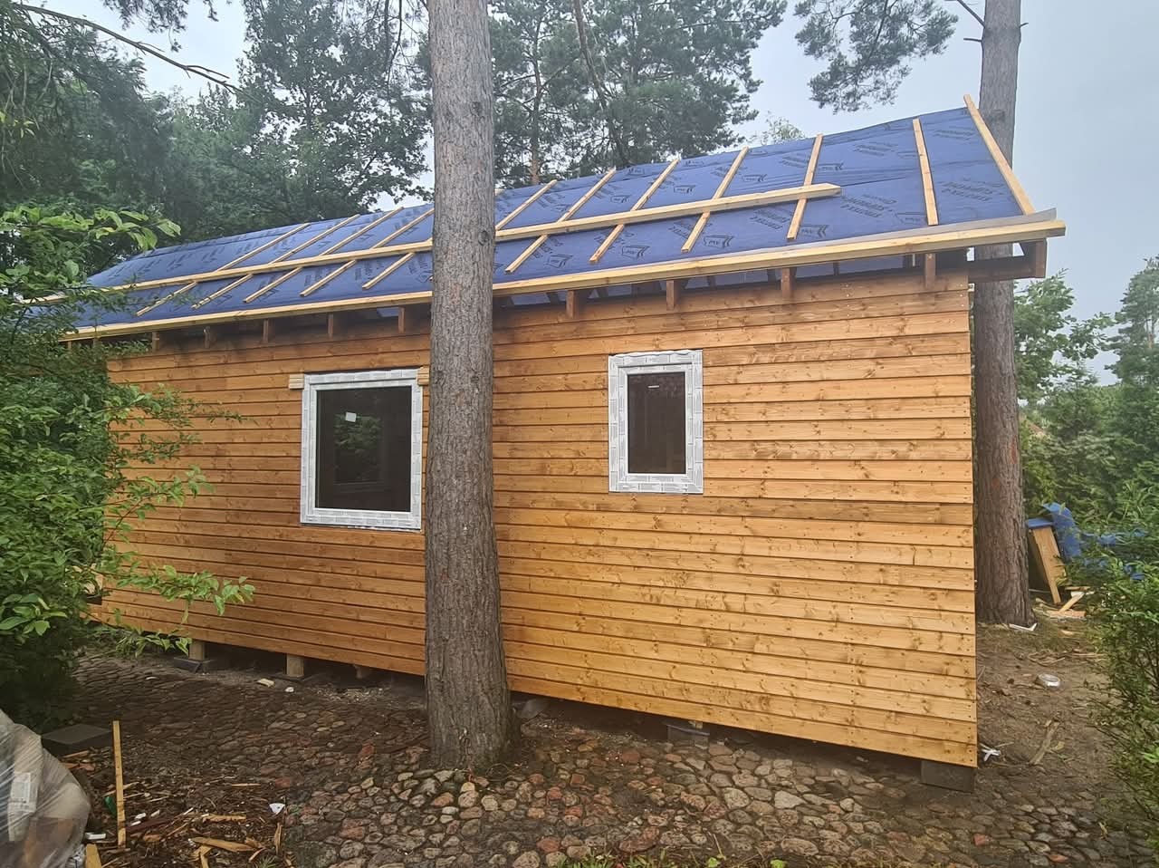 Drewniany dom szkieletowy w trakcie budowy, widoczne okna i wstępne krycie dachu z niebieską membraną. Drzewo rosnące blisko ściany. Kamienna podstawa.