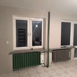 Marcin Adamski - Pokój po remoncie: dwa okna z grzejnikami (zielony i szary), ściany w odcieniach szarości, jedna z wzorzystą tapetą. Widoczne rury grzewcze.