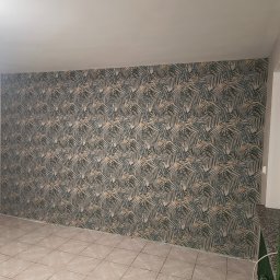 Marcin Adamski - Pokój po tapetowaniu: ściana w liście palmowe, beżowe płytki podłogowe, widoczny fragment okna z zielonym grzejnikiem. Jasne wnętrze po remoncie.