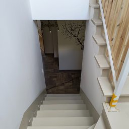 Marcin Adamski - Widok z góry na jasne schody z drewnianą balustradą i oświetleniem punktowym w ścianie. W tle widoczny fragment podłogi i dekoracja ścienna z motywem drzewa.