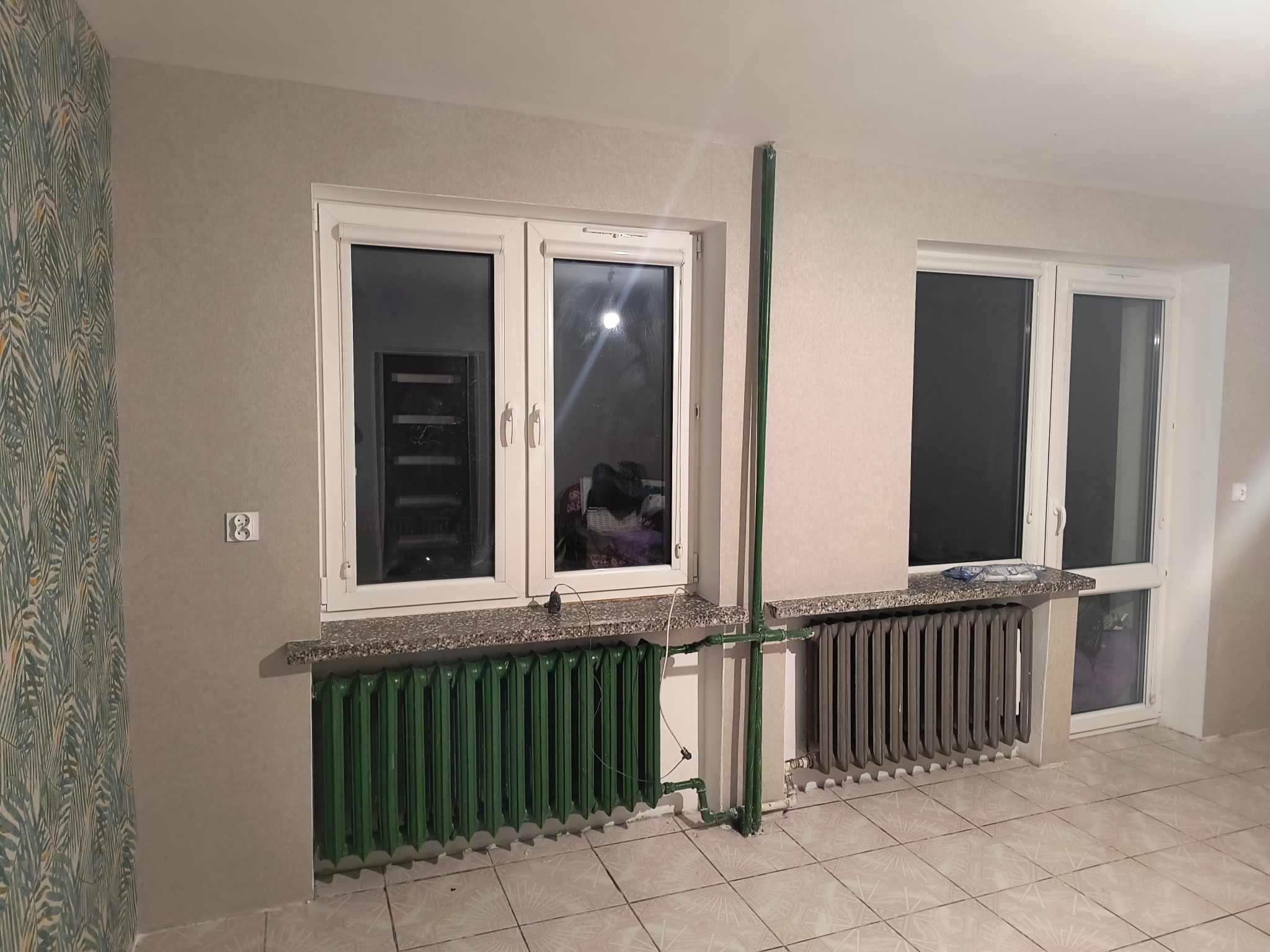 Pokój po remoncie: dwa okna z grzejnikami (zielony i szary), ściany w odcieniach szarości, jedna z wzorzystą tapetą. Widoczne rury grzewcze.