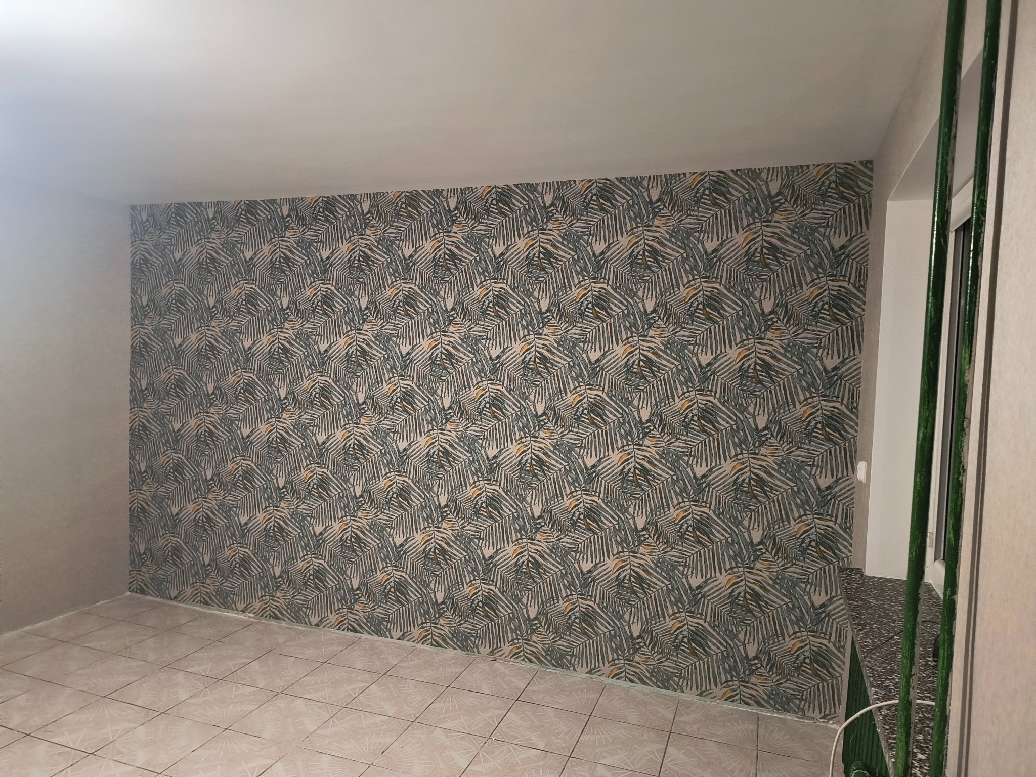 Pokój po tapetowaniu: ściana w liście palmowe, beżowe płytki podłogowe, widoczny fragment okna z zielonym grzejnikiem. Jasne wnętrze po remoncie.