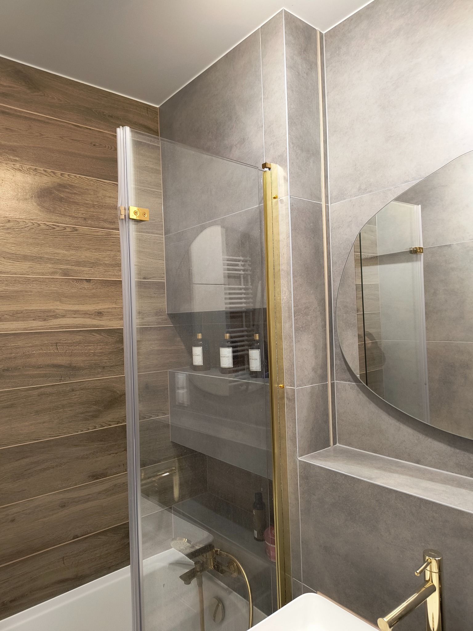 Nowoczesna łazienka z drewnopodobnymi i szarymi płytkami, kabiną prysznicową ze złotymi detalami i okrągłym lustrem. Minimalistyczny design i eleganckie wykończenie.