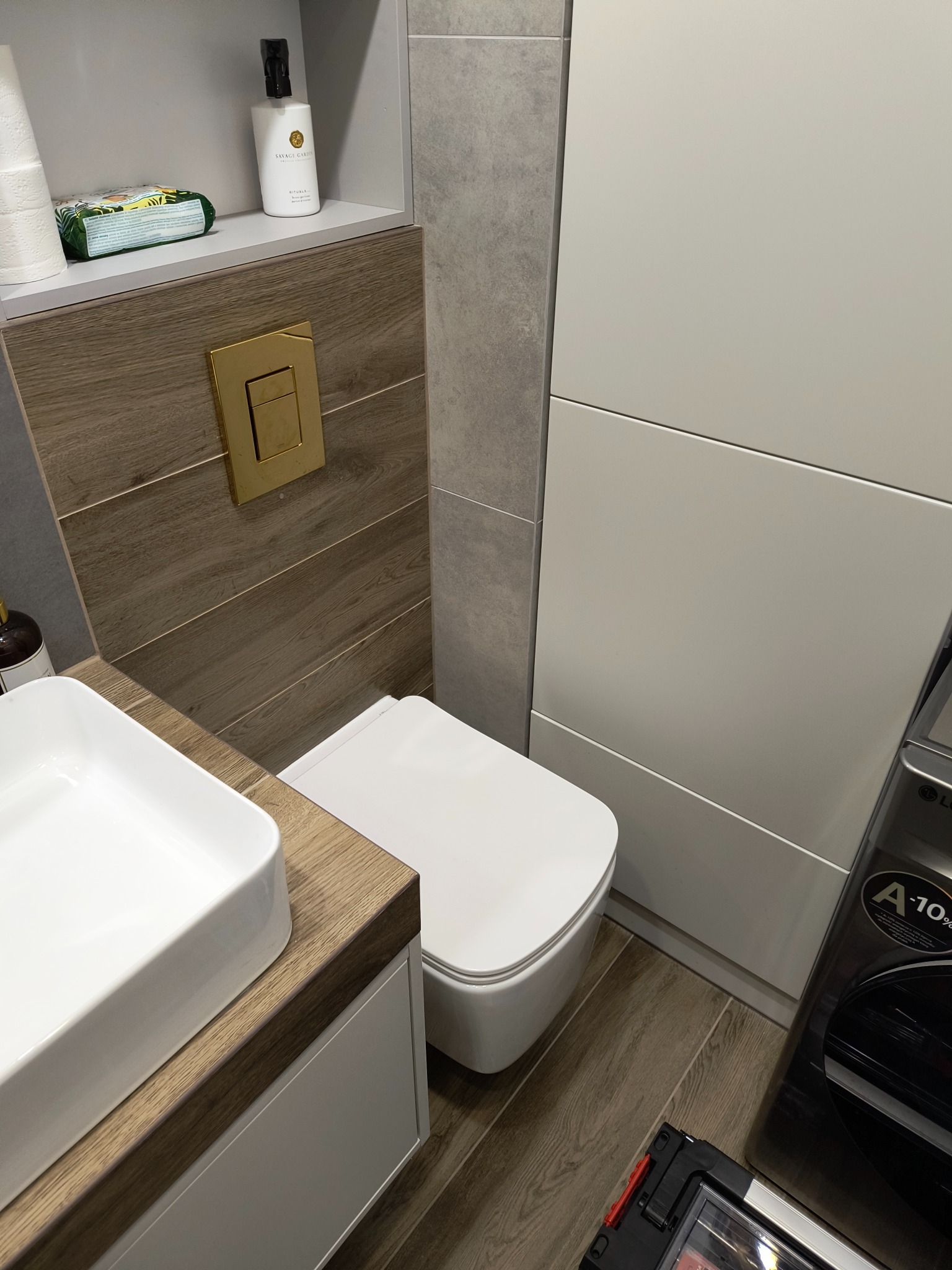 Nowoczesna łazienka z drewnopodobnymi płytkami, białą ceramiką i złotym akcentem w postaci przycisku spłuczki. Umywalka nablatowa i zabudowa pralki.