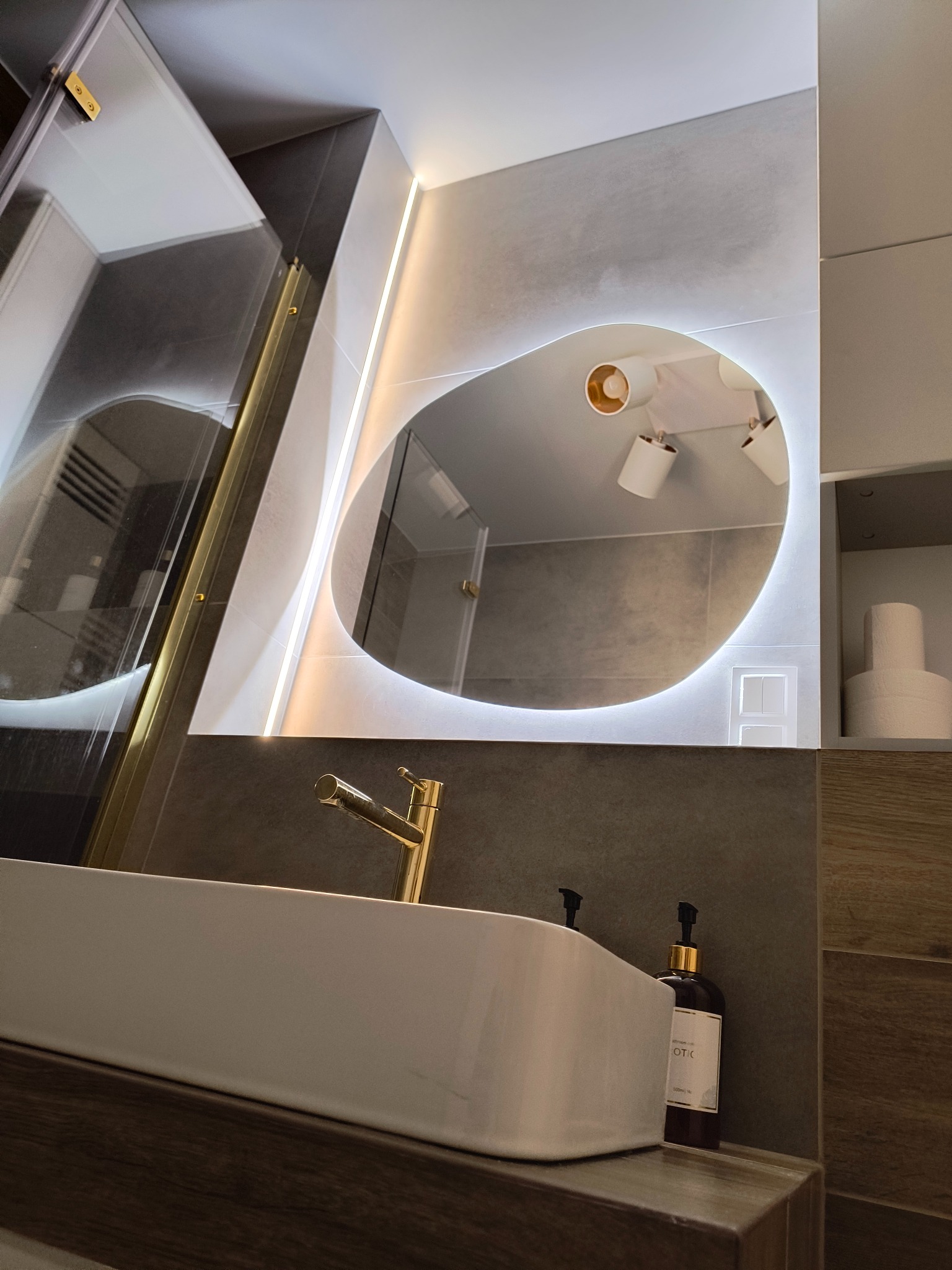 Nowoczesna łazienka: biała umywalka, złota bateria, lustro z podświetleniem LED i kabina prysznicowa z elementami w kolorze złotym. Minimalistyczny design.