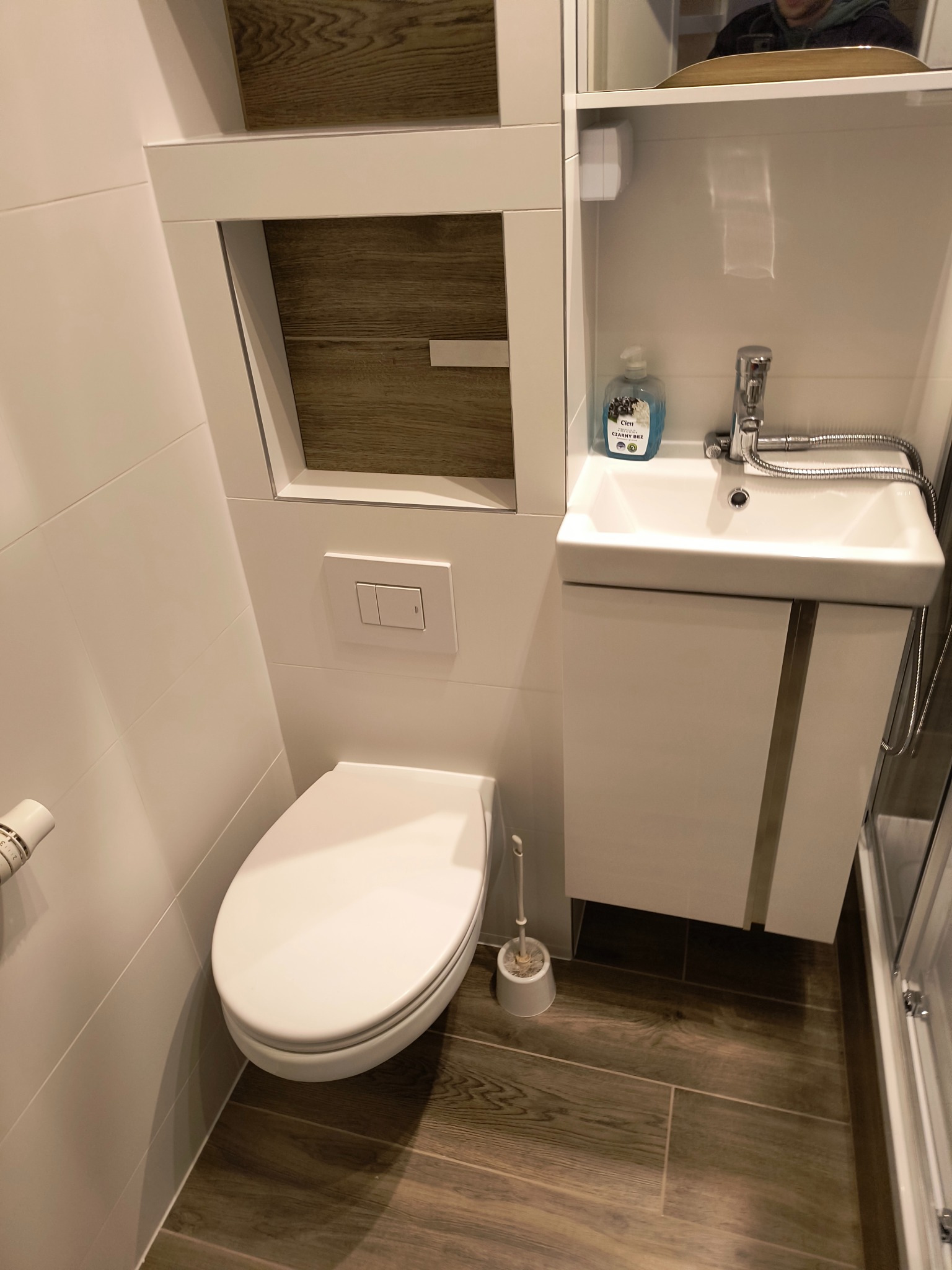 Nowoczesna łazienka z podwieszaną toaletą, umywalką z szafką i drewnopodobną podłogą. Białe płytki ścienne i minimalistyczny design. Widoczny dozownik mydła.