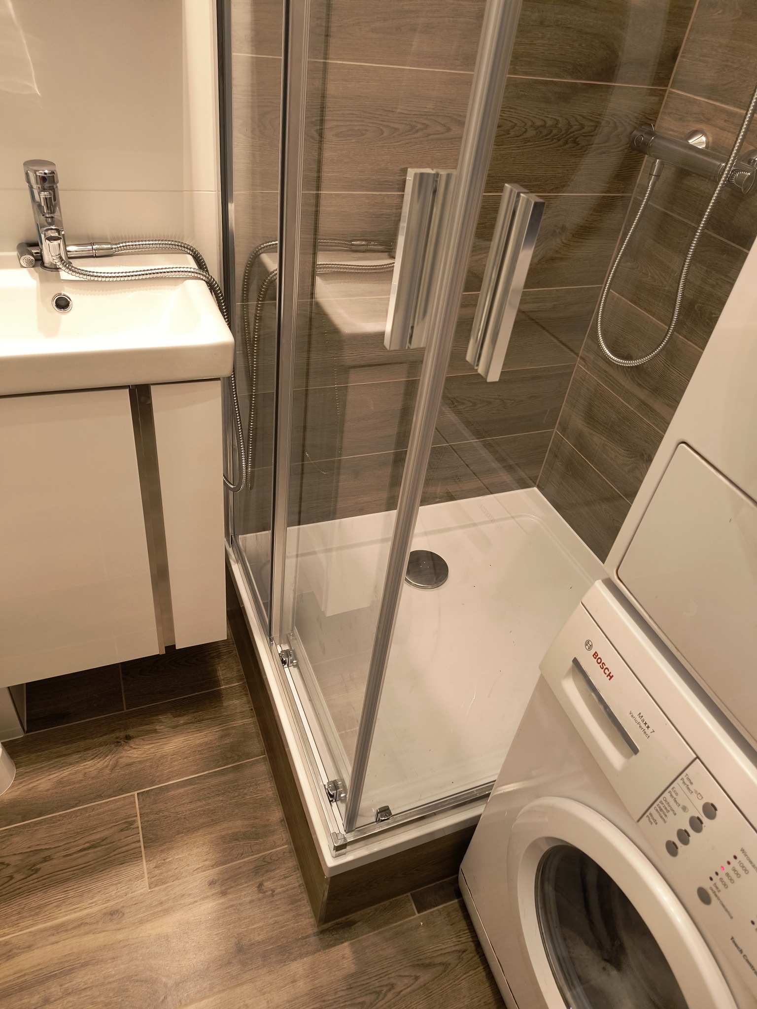 Nowoczesna łazienka z kabiną prysznicową, umywalką z szafką i pralką Bosch. Płytki imitujące drewno na podłodze i ścianach. Chromowana armatura.