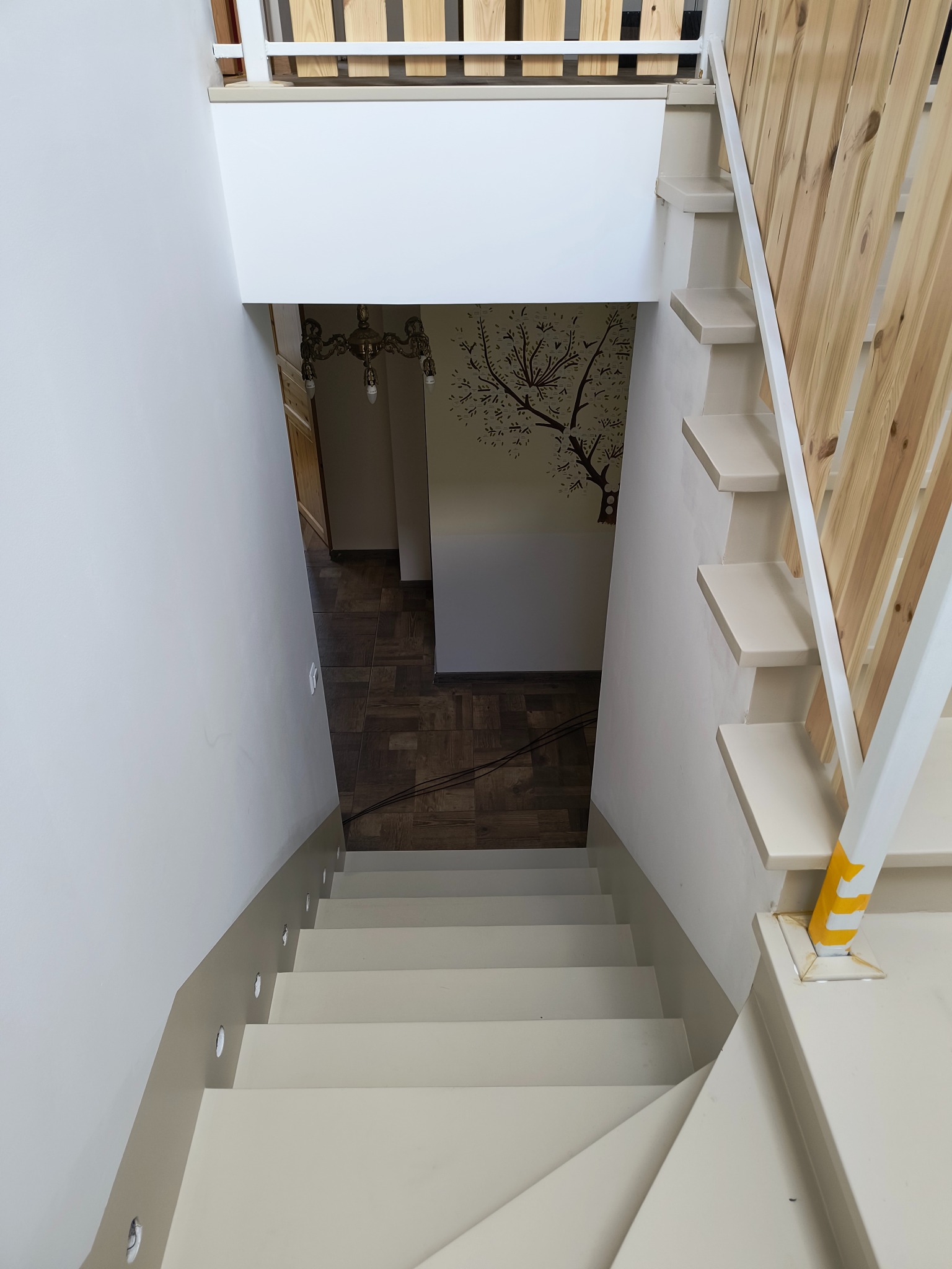Widok z góry na jasne schody z drewnianą balustradą i oświetleniem punktowym w ścianie. W tle widoczny fragment podłogi i dekoracja ścienna z motywem drzewa.