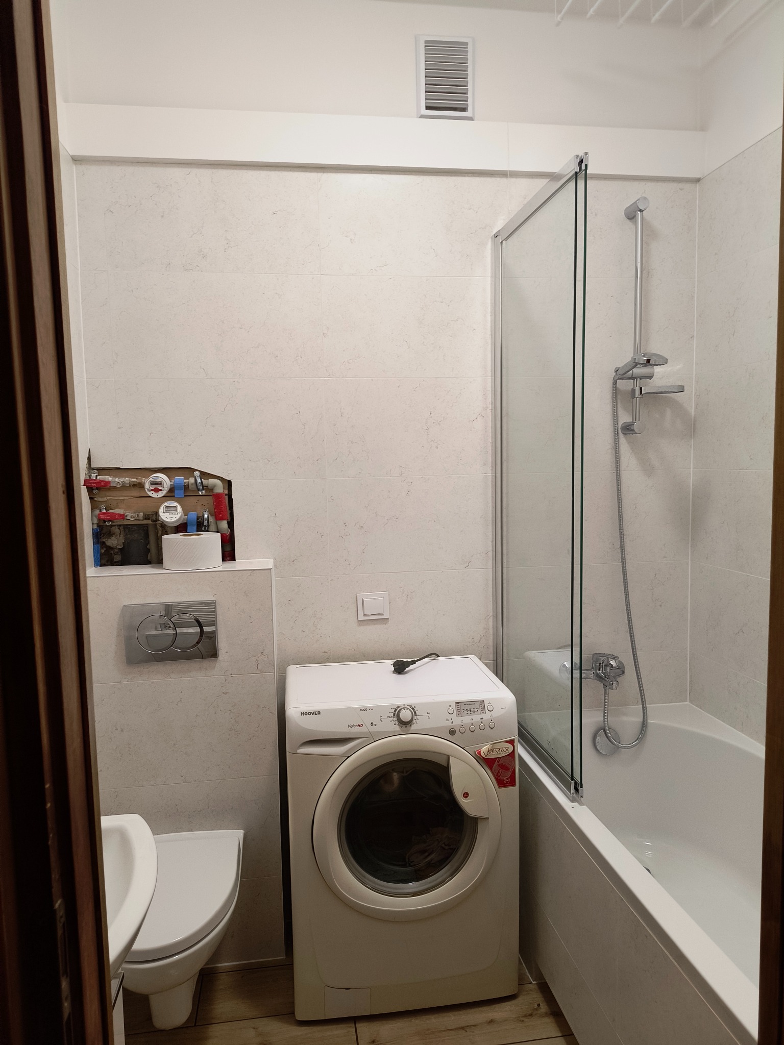 Wyremontowana łazienka z pralką, kabiną prysznicową i toaletą. Widoczne płytki w jasnych odcieniach, drewniane wykończenia drzwi i instalacja wodna nad toaletą.