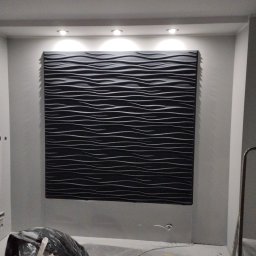 Gładzie Nowacki - Czarny panel dekoracyjny 3D z poziomymi falami na ścianie w szarym pokoju, oświetlony punktowo. Widoczne ślady prac remontowych na podłodze i drabina.