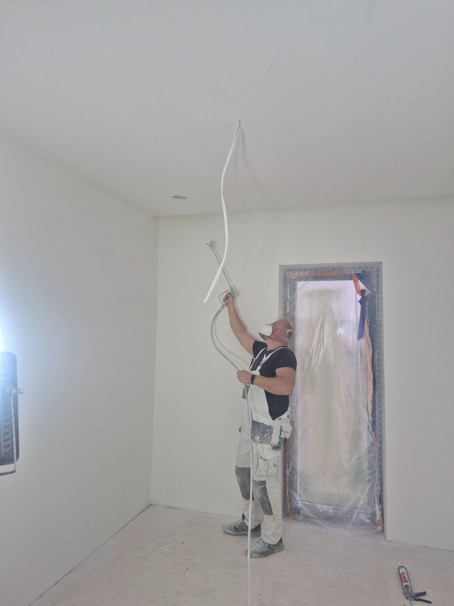 Pracownik w kombinezonie i masce maluje sufit agregatem natryskowym w jasnym pomieszczeniu. Zabezpieczone drzwi folią. Widoczne narzędzia i oświetlenie.