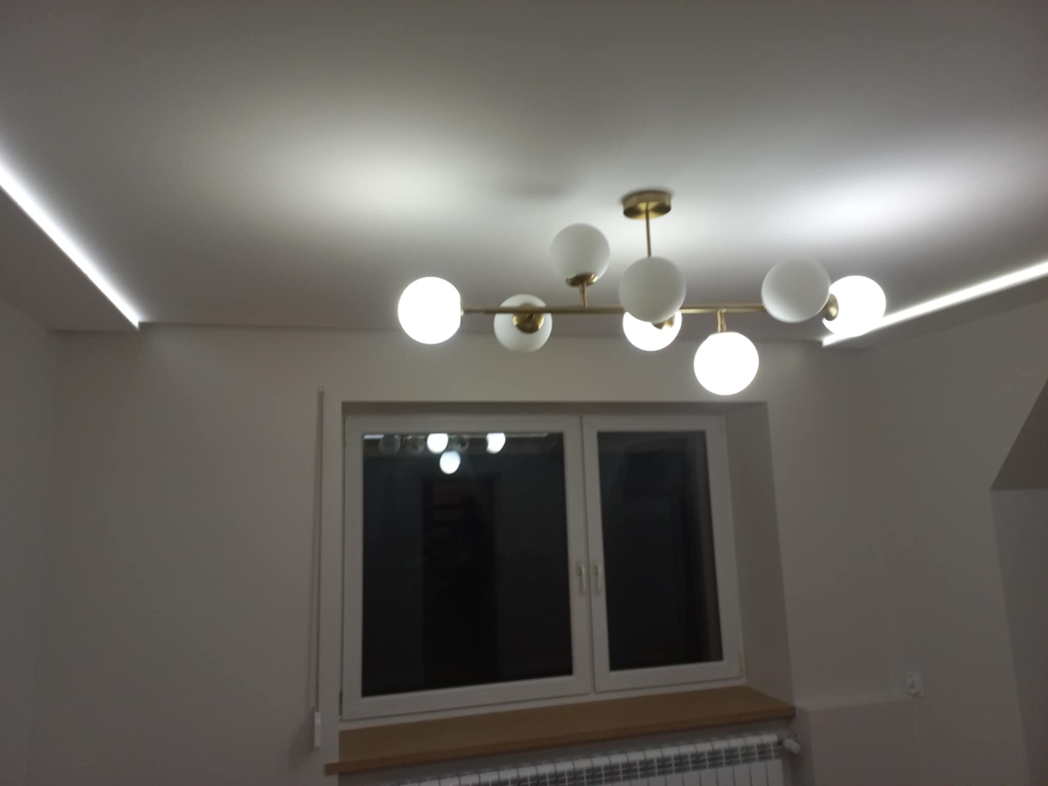 Nowoczesne oświetlenie sufitowe z ledami wzdłuż krawędzi sufitu i lampą kulistą. Świeżo pomalowane ściany w neutralnym odcieniu. Widok na okno.