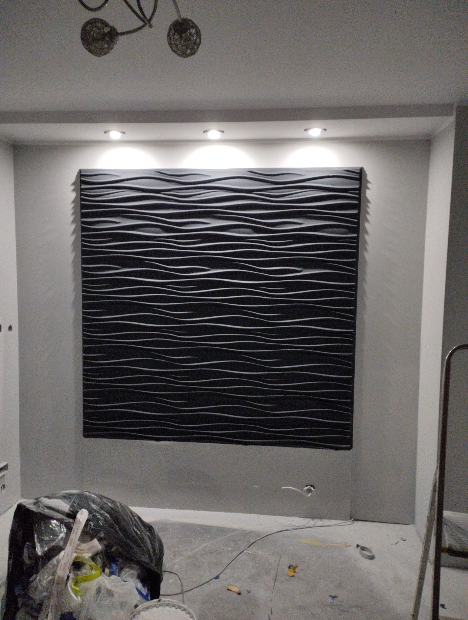 Czarny panel dekoracyjny 3D z poziomymi falami na ścianie w szarym pokoju, oświetlony punktowo. Widoczne ślady prac remontowych na podłodze i drabina.