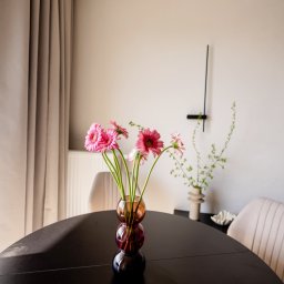 Casa Decorada - Stylowy stół z ciemnego drewna, na nim wazon z różowymi gerberami. W tle minimalistyczny zegar ścienny i beżowe zasłony. Subtelna dekoracja wnętrza.