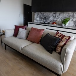Casa Decorada - Jasna sofa z poduszkami w salonie z aneksem kuchennym. Widoczny marmurowy blat, drewniana podłoga i abstrakcyjny obraz. Neutralna kolorystyka, nowoczesny design.