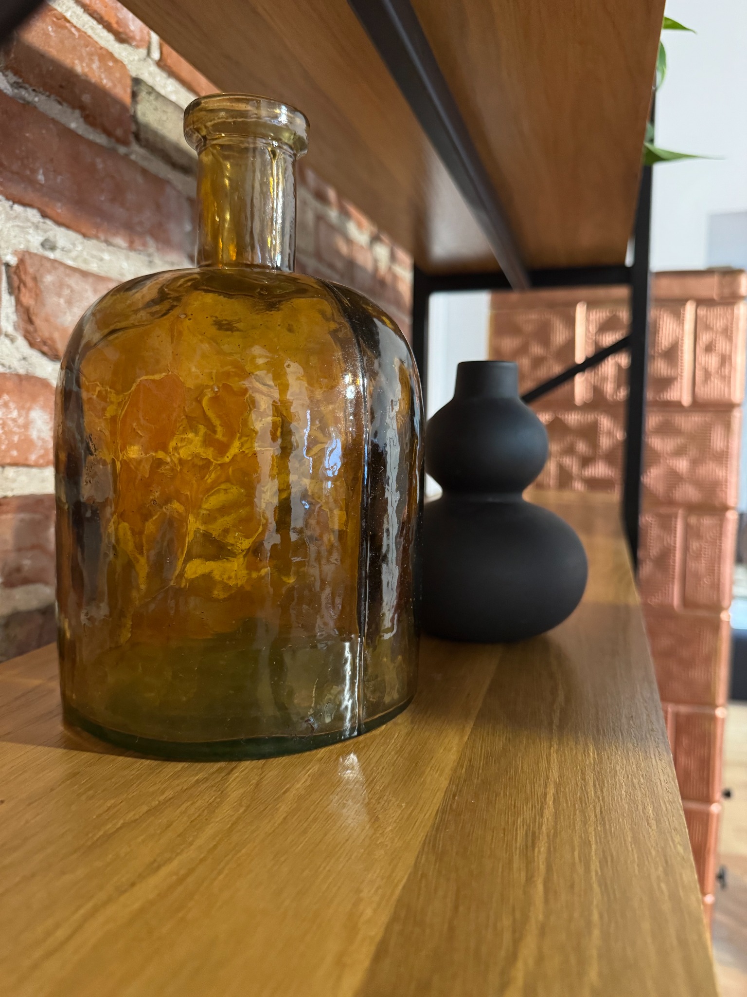 Dekoracyjna, bursztynowa butelka i czarny wazon na drewnianej półce z ceglanym tłem. Element wystroju wnętrza w stylu vintage.