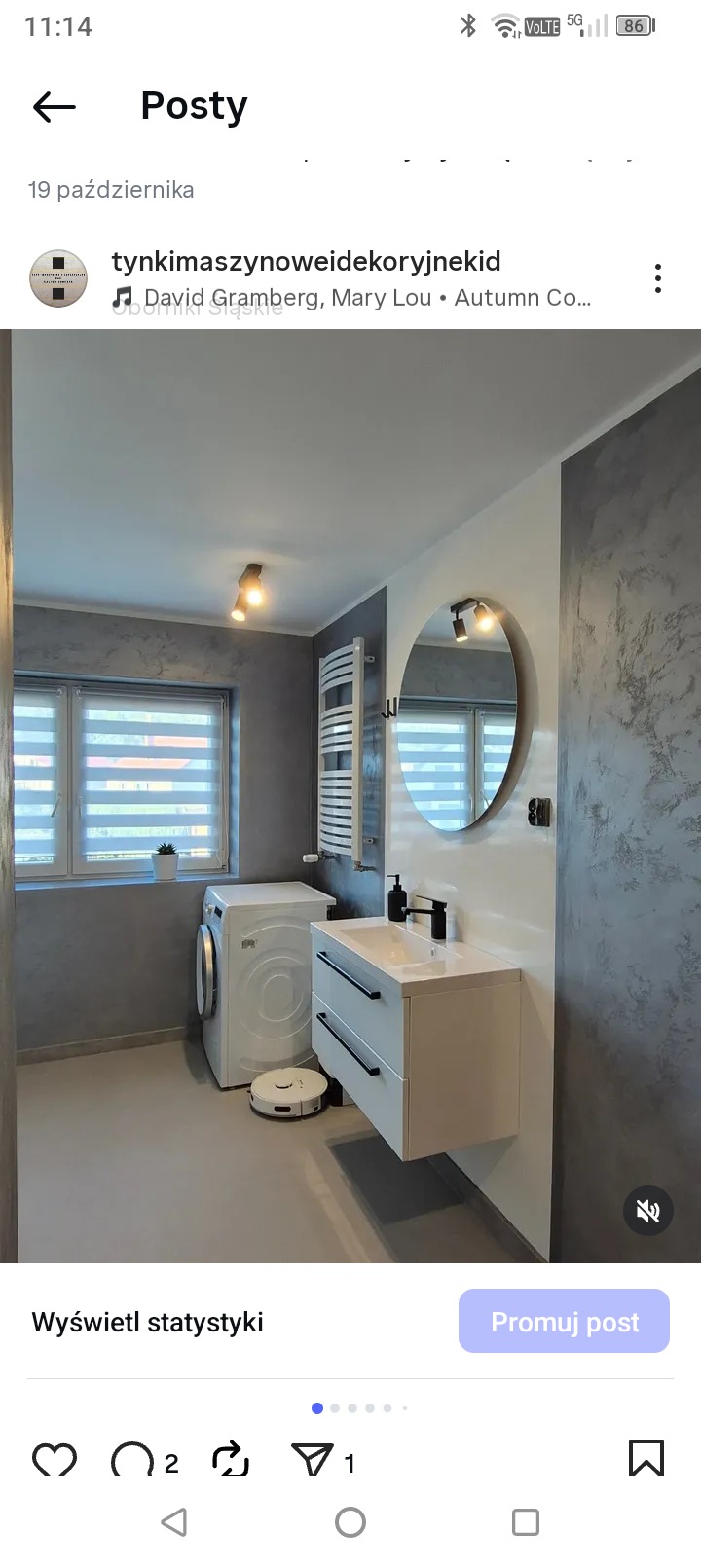 Szara łazienka z pralką, okrągłym lustrem i umywalką z szafką. Ściany wykończone dekoracyjnym tynkiem. Widoczny robot sprzątający. Nowoczesny design.