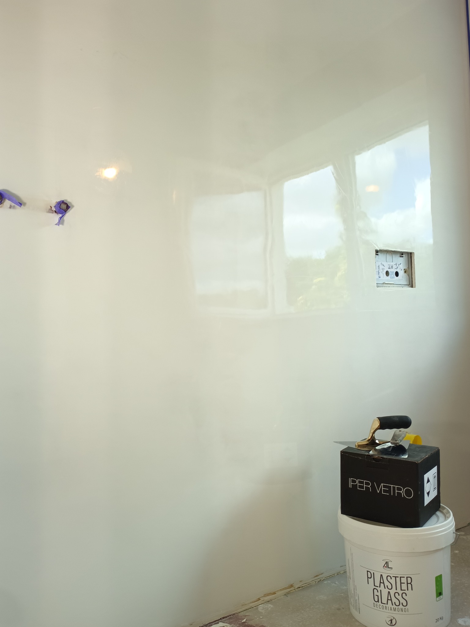 Ściana po szpachlowaniu z efektem lustra, widoczne puszki elektryczne zabezpieczone taśmą, wiadro z gładzią i szpachelka. Jasne wnętrze, światło dzienne.