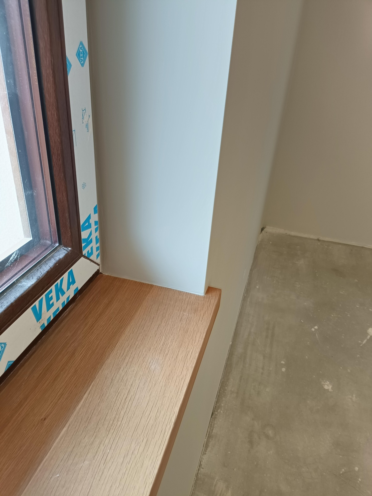 Zamontowany drewniany parapet okienny w odcieniach brązu, białe ściany i surowa podłoga. Widoczne okno z brązową ramą i folią ochronną.