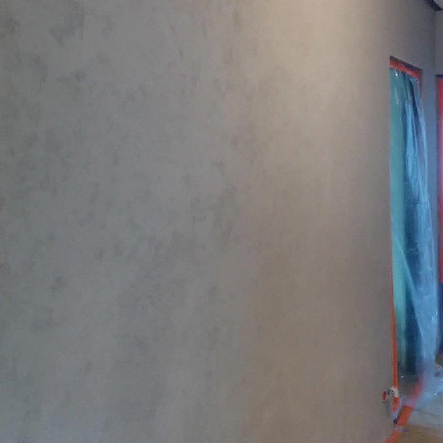 Ściana z efektem dekoracyjnym w odcieniach szarości, zabezpieczone folią drzwi w tle. Widoczne gniazdko elektryczne przy podłodze.