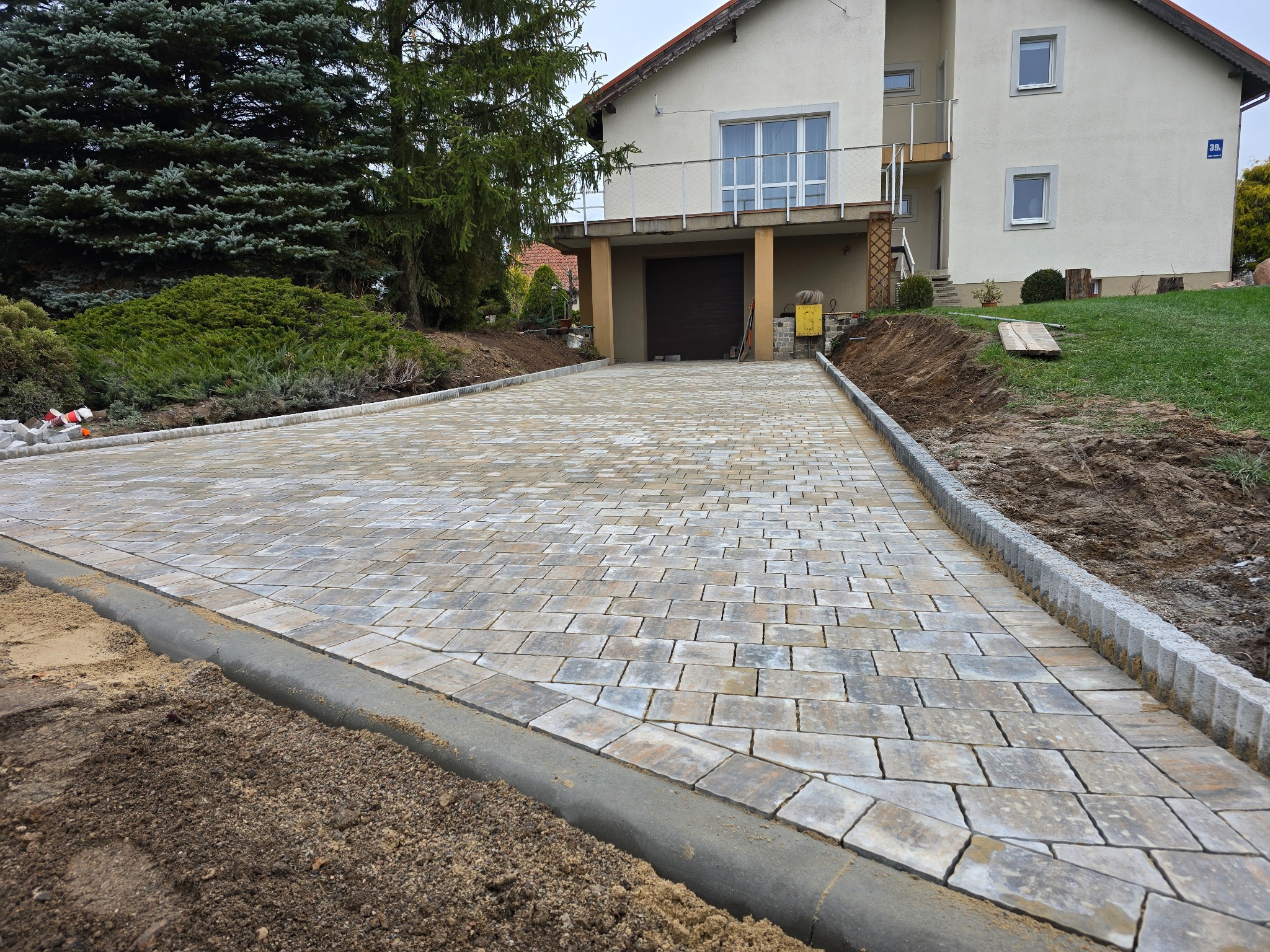 Nowa, jasna kostka brukowa prowadząca do domu z garażem, obrzeża betonowe, świeżo obsypane ziemią. Widok na dom i zieleń w tle.