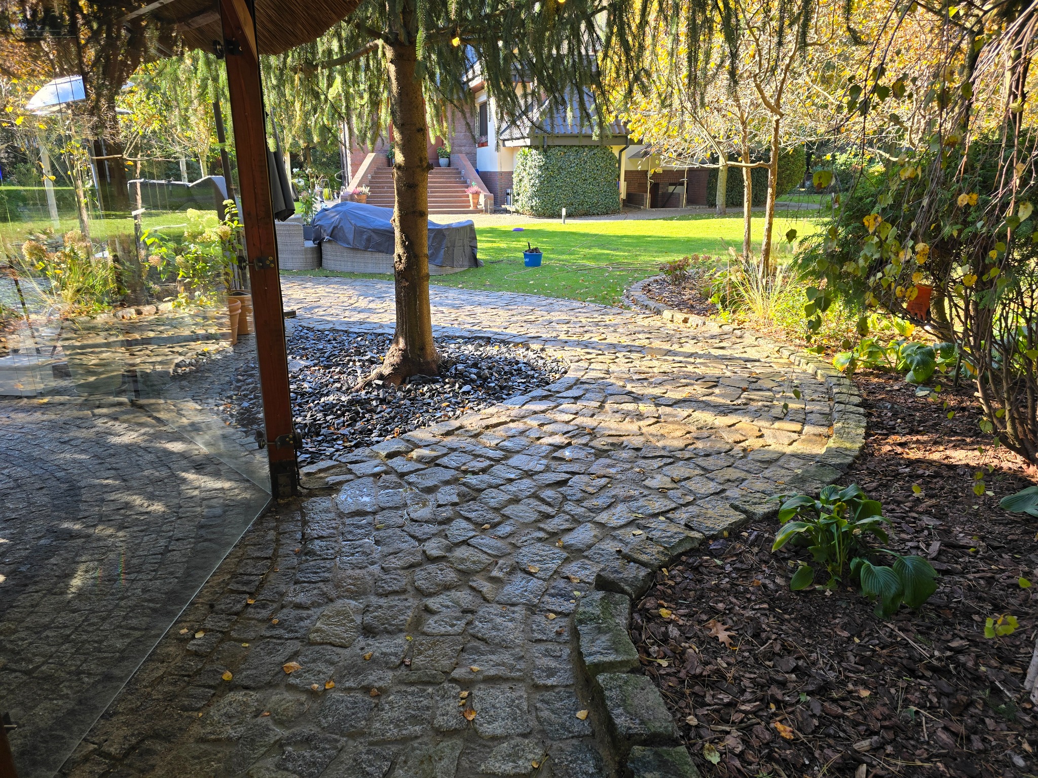 Kręta ścieżka z kostki brukowej prowadząca przez ogród z drzewem pośrodku, czarnymi kamieniami i zielonym trawnikiem w tle, widziana z zadaszonego tarasu.