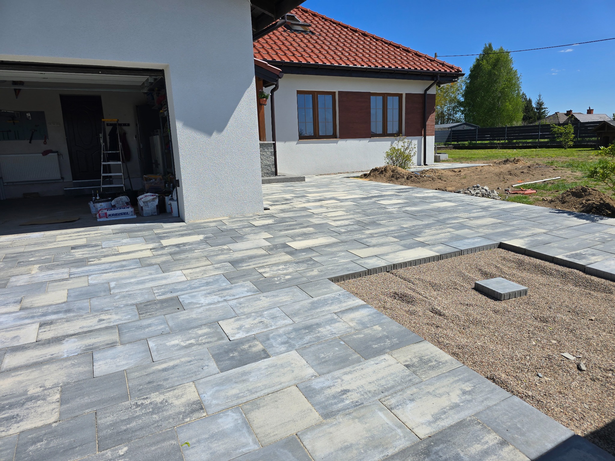 Ułożona kostka brukowa w odcieniach szarości prowadząca do domu z czerwoną dachówką. Fragment terenu przygotowany pod dalsze prace brukarskie z podsypką.