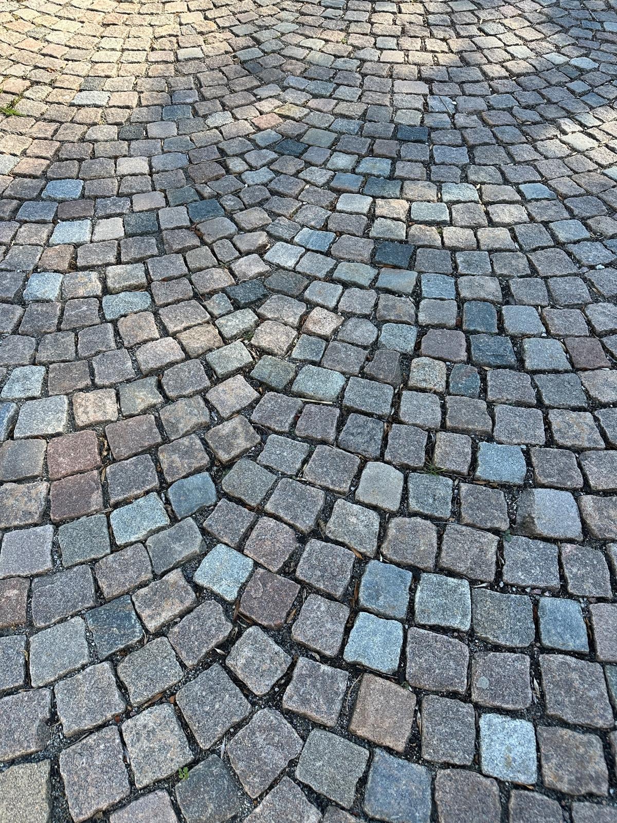 Ułożona kostka brukowa w odcieniach szarości i brązu, tworząca wzór łuku. Widoczne spoiny i delikatne cienie podkreślają fakturę kamienia.