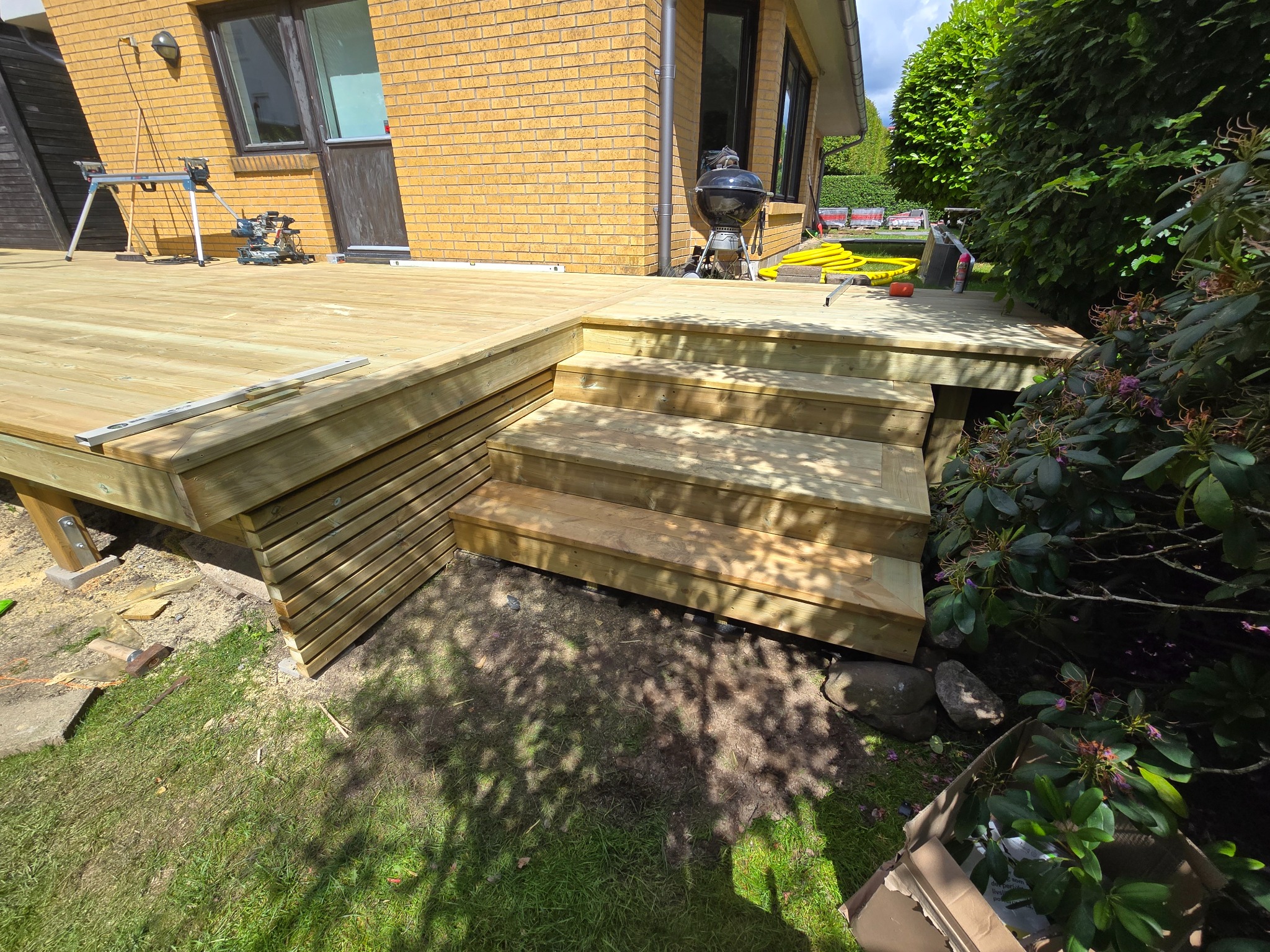 Drewniany taras z schodami w Purdzie, częściowo ukończony, z widocznymi narzędziami i grillem. Jasne drewno, zielony trawnik i krzewy w tle.