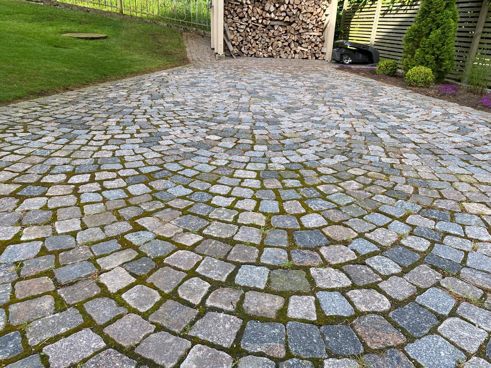 Ułożona z kamiennej kostki brukowej, półkolista ścieżka z delikatnym mchem między kamieniami. W tle trawnik, drewno na opał i robot koszący.