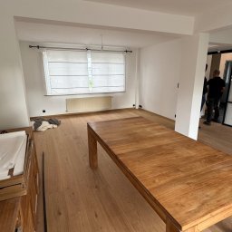 Patryk Leonkiewicz - Jasne, świeżo pomalowane wnętrze z drewnianą podłogą i stołem. Widoczne okno z roletą i fragment instalacji oświetleniowej. W tle sylwetki osób.