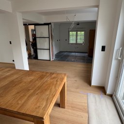 Patryk Leonkiewicz - Wnętrze w trakcie remontu: drewniany stół na pierwszym planie, w tle widoczne płytki podłogowe i przesuwne drzwi. Widoczne przygotowania do prac wykończeniowych.