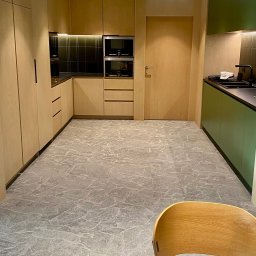 Patryk Leonkiewicz - Nowoczesna kuchnia z drewnianymi szafkami i szarym marmurkowym dywanem. Zielone akcenty w zabudowie kuchennej. Widoczny fragment krzesła w prawym dolnym rogu.