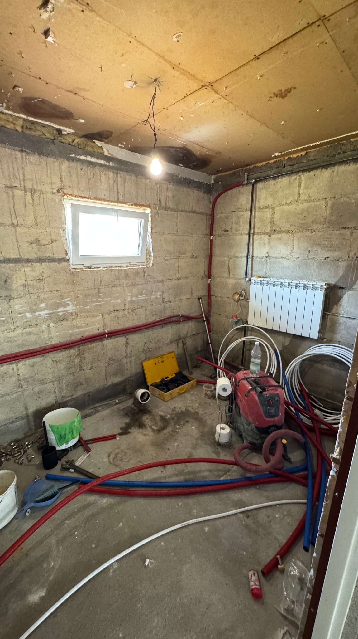 Surowy pokój z instalacją grzewczą: grzejnik, rury czerwone i niebieskie, narzędzia w żółtej skrzynce. Widoczne surowe ściany z betonu komórkowego i prowizoryczne oświetlenie.
