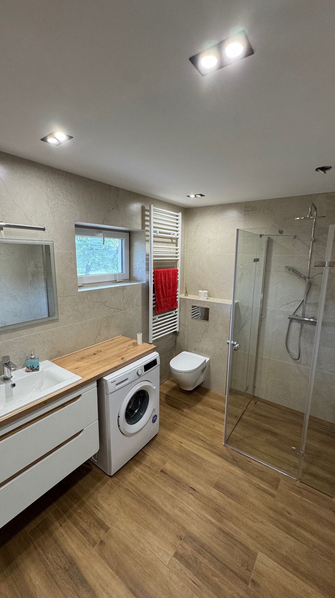 Jasna łazienka z drewnianą podłogą i blatem, kabiną prysznicową, pralką i grzejnikiem drabinkowym z czerwonym ręcznikiem. Minimalistyczny design, neutralna kolorystyka.