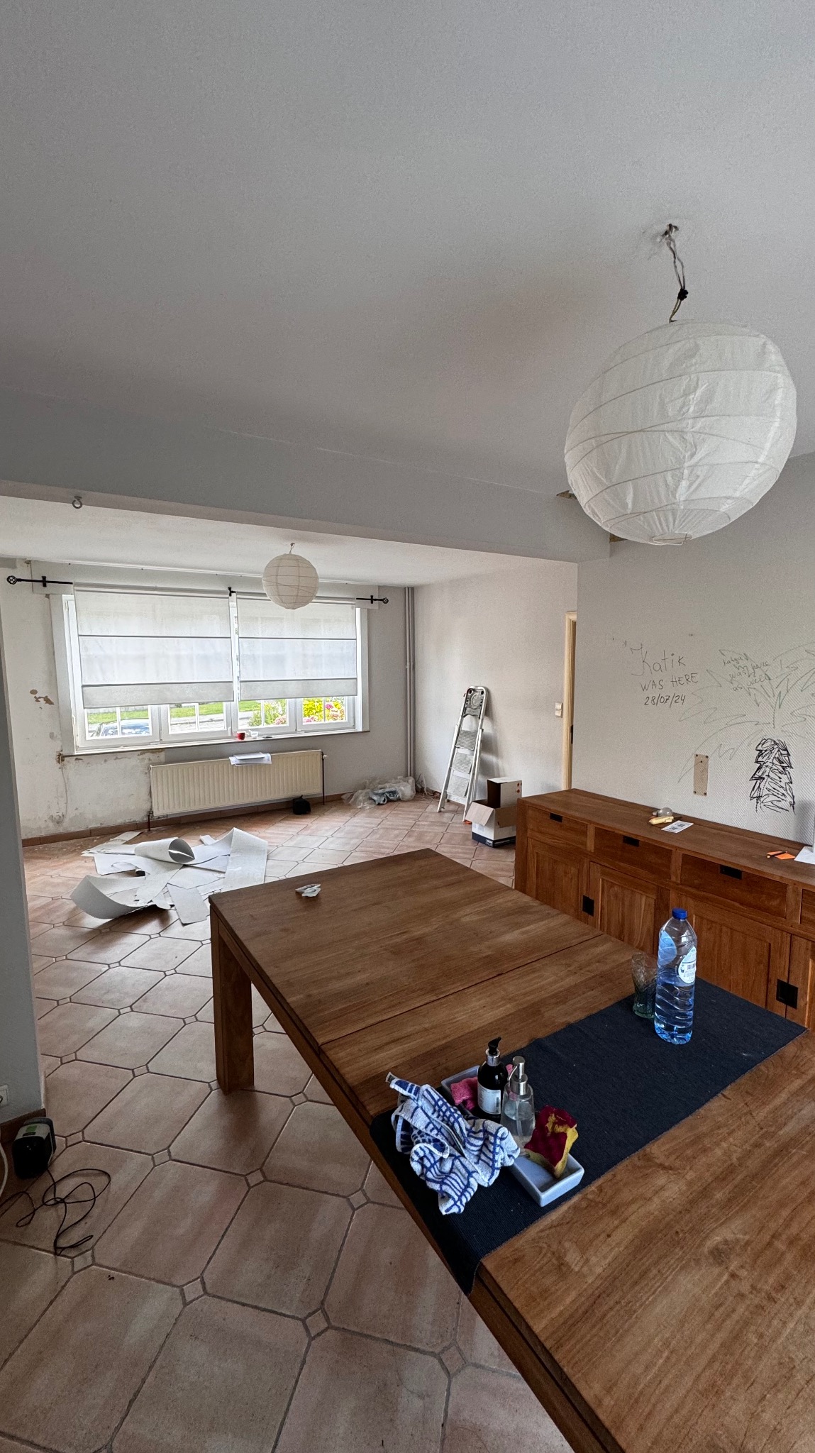 Pokój w trakcie remontu: zerwane tapety, drabina oparta o ścianę z dziecięcymi rysunkami, duży drewniany stół na pierwszym planie. Widoczne przygotowania do malowania.