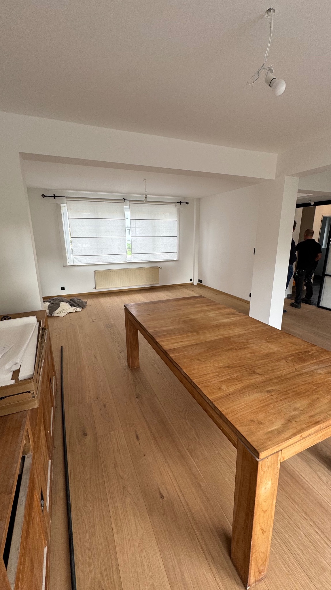 Jasne, świeżo pomalowane wnętrze z drewnianą podłogą i stołem. Widoczne okno z roletą i fragment instalacji oświetleniowej. W tle sylwetki osób.