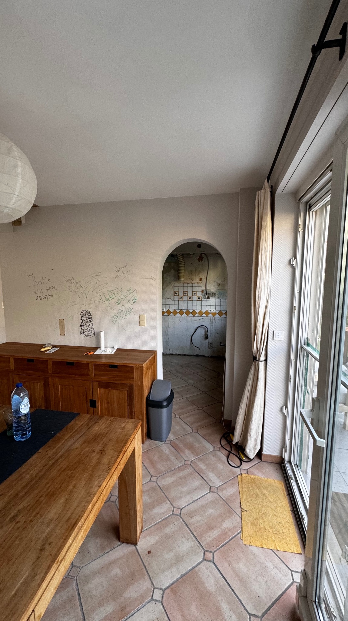 Wnętrze w trakcie remontu: widoczna ściana z napisami, drewniane meble, terakota na podłodze i przejście do zdemontowanej kuchni. Widok na okno balkonowe.