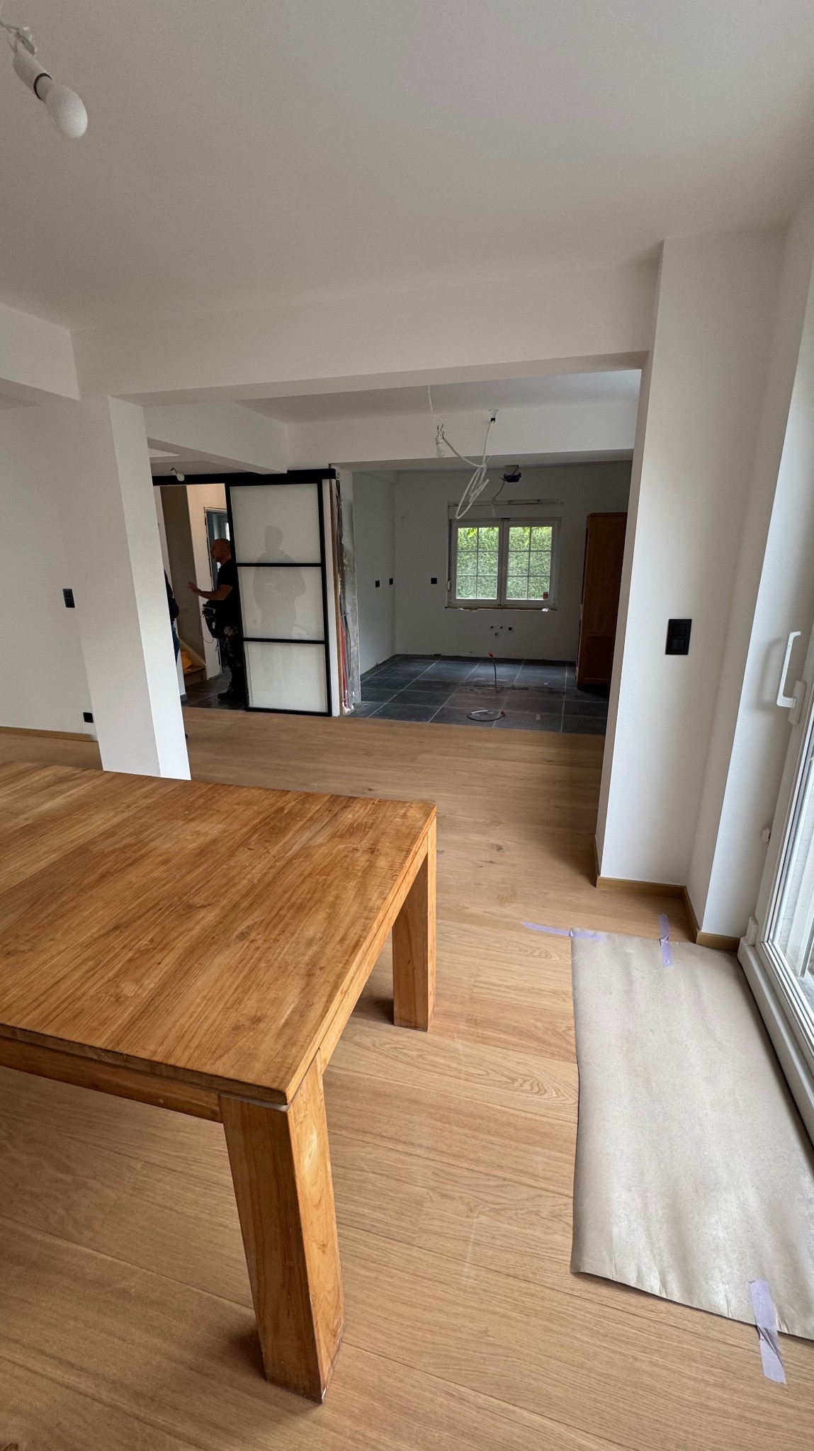 Wnętrze w trakcie remontu: drewniany stół na pierwszym planie, w tle widoczne płytki podłogowe i przesuwne drzwi. Widoczne przygotowania do prac wykończeniowych.