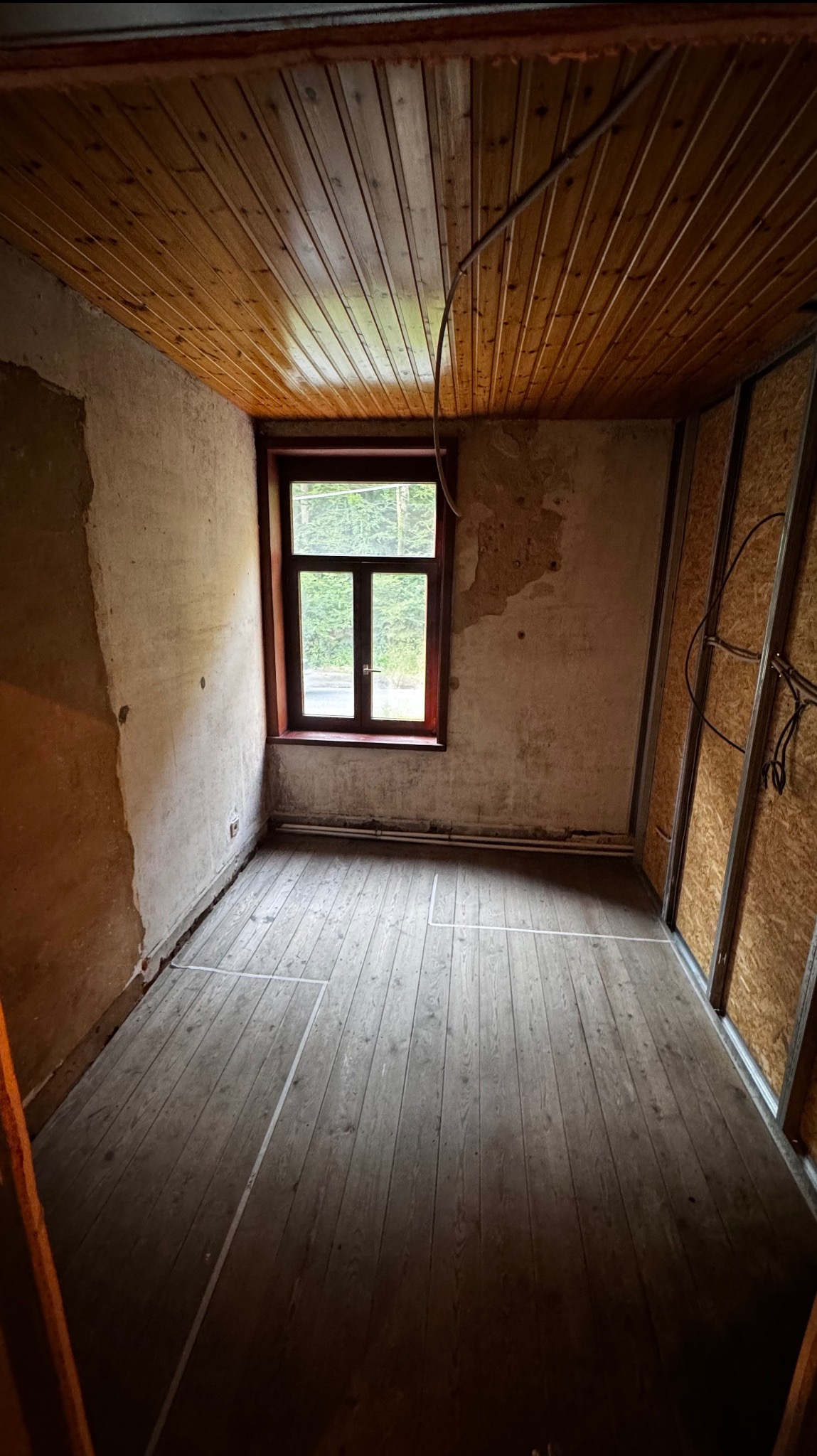 Pokój przed remontem: drewniana podłoga z taśmami, ściany z ubytkami tynku, drewniany sufit i okno. Widoczne przygotowania do prac wykończeniowych.